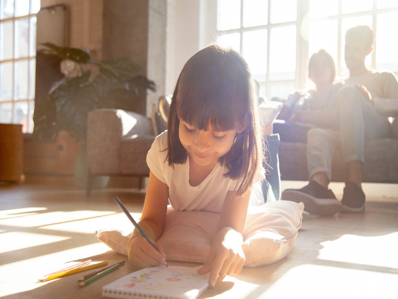 Young, happy girl lays on floor and draws while family relaxes on couch in home that uses a YORK Ultraviolet Air Treatment System.
